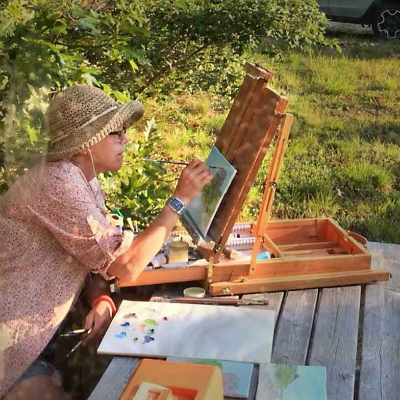 Artist painting en plein air, seated at a picnic table, captured unawares.